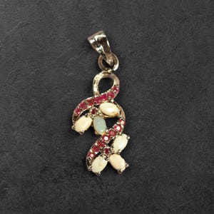 May include: A silver-toned pendant featuring a swirling, ribbon-like design set with multiple oval-shaped white opals and small, round, deep red gemstones. The pendant includes a polished metal bail at the top for attachment to a necklace chain.