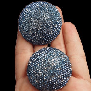 May include: A pair of circular, dome-shaped earrings featuring a dense pavé setting of small, sparkling blue and silver crystals. The earrings have a textured, glittery surface that catches the light, creating a multi-tonal blue appearance.