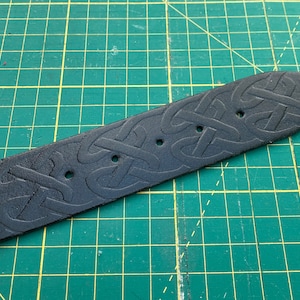 Puede incluir: Una correa de cinturón de cuero negro con un patrón de nudo celta en relieve. La correa tiene tres agujeros espaciados para el ajuste y un extremo redondeado. El fondo es una esterilla de corte verde.