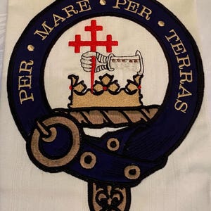 May include: A blue and gold embroidered crest with a crown, a hand holding a sword, and a cross. The text around the crest reads "Per Mare Per Terras".