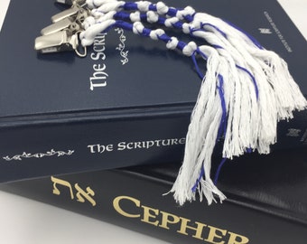 May include: Three white and blue braided cord bookmarks with silver metal clips attached to the top. The bookmarks feature knotted patterns and long, loose fringe ends, resting on top of a dark blue book titled The Scriptures.