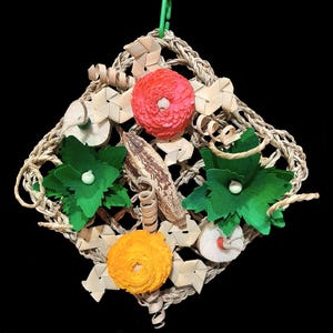May include: A diamond-shaped bird toy made from woven material, with a green hanging hook. It has decorative elements including red and yellow flower shapes, green leaf shapes, and natural wood-coloured pieces. The toy is designed for avian enrichment.
