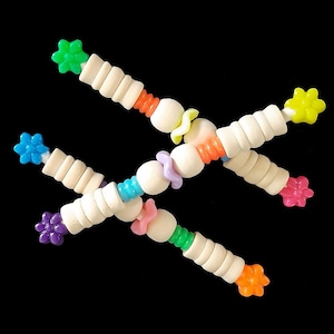 May include: Four colourful wooden toy sticks with flower-shaped ends and various coloured beads. The sticks are arranged in a crisscross pattern against a black background. The colours include green, blue, pink, purple, yellow, and orange.