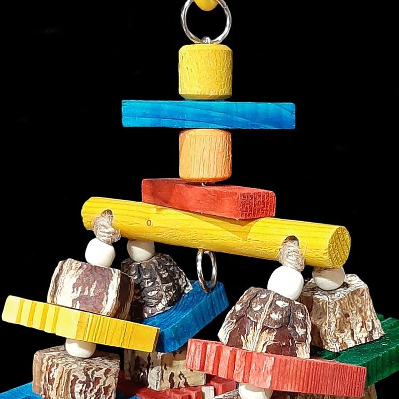 Layers of soft mahogany pod chunks, brightly colored notched pine busy beaver slices and small wood snap beads flowing under a wood dowel base on jute cord. Hangs on a short length of nickel plated chain with more wood pieces. Designed for intermediate to medium sized birds who like softer textures (cockatiels, small conures, quakers, caiques, greys, etc). Measures approx 5" by 10" including link.
