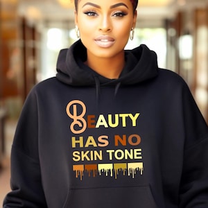 Può includere: Una felpa nera con cappuccio che presenta il testo 'BEAUTY HAS NO SKIN TONE' stampato in lettere gialle e marroni sul petto. Sotto il testo c'è un grafico di cinque rettangoli orizzontali in varie tonalità di pelle con un effetto gocciolante.
