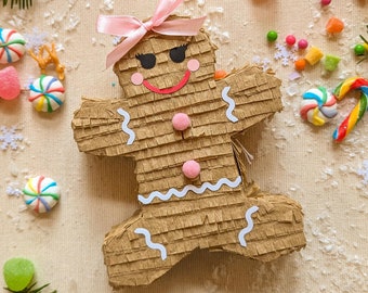 May include: A brown gingerbread man piñata with a pink bow, pink cheeks, and a red smile. The piñata is surrounded by colorful candies, candy canes, and sprigs of evergreen. Festive holiday decor.