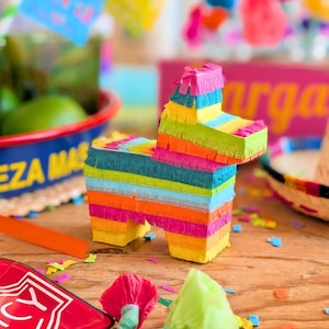 May include: A colorful, donkey-shaped piñata made of layered paper in shades of yellow, orange, pink, blue, green, and yellow. The piñata is decorated with fringed paper and sits on a wooden surface, with party decorations in the background.