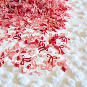 May include: A scattering of red and pink paper confetti and bow-shaped cutouts on a white, textured surface. The confetti includes small rectangular pieces and bow shapes in various shades of red and pink, creating a festive and decorative display.