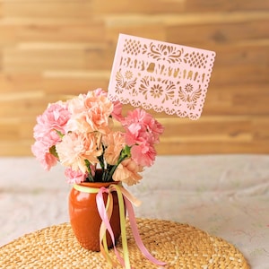 Cielito Lindo Centerpiece Flag - Custom Papel Picado by Lula Flora. Close-up of a personalized baby shower mini papel picado table flag in blush pink cardstock with floral cutouts and scalloped border, mounted on a pick. Set in a bouquet of pink and peach carnations in a small clay-style vase tied with pink and sage ribbons, on a woven placemat with a warm wood background. Mexican-inspired centerpiece décor; customizable with baby’s name or message; ideal for tables, dessert bar, or gift table.