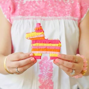 May include: A colorful donkey piñata made of layered pink, orange, and yellow crepe paper. The piñata is held in front of a white blouse with pink embroidery. The piñata is a festive decoration for parties and celebrations.