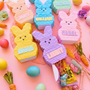 May include: A collection of colorful Easter bunny piñatas in shades of orange, blue, green, pink, yellow, and purple, each with a name on the front. The piñatas are surrounded by Easter eggs, candies, and decorative carrots on a pink surface.