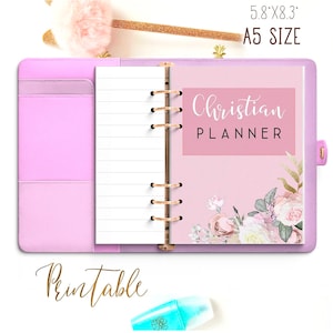 Op de afbeelding: Een roze A5-planner met een bloemenontwerp en de tekst "Christian Planner" op de omslag. De planner is open op een blanco pagina met gestreept papier.