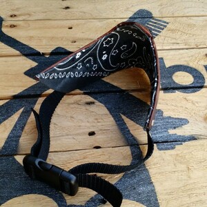 May include: A black bandana with a white paisley pattern is attached to a brown leather strap with a black adjustable buckle. The strap is on a wooden surface.