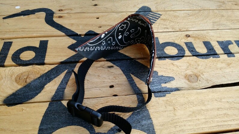 May include: A black bandana with a white paisley pattern is attached to a brown leather strap with a black adjustable buckle. The strap is on a wooden surface.