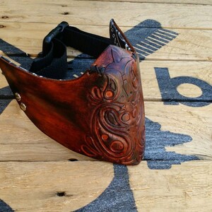 May include: A brown leather mask with intricate floral carvings. The mask has an adjustable strap and is secured with metal rivets.