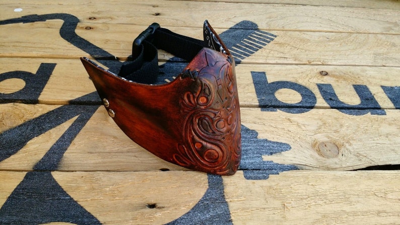 May include: A brown leather mask with intricate floral carvings. The mask has an adjustable strap and is secured with metal rivets.