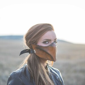 May include: A woman wearing a brown leather mask with a black strap. The mask has a pointed beak and is covering the lower half of her face. She is wearing a black leather jacket and has long brown hair.