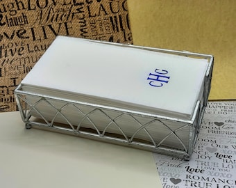 May include: A silver metal napkin holder with a stack of white linen napkins. The napkins have a blue monogram that reads "CHG".