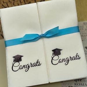 May include: Two white cards tied together with a blue ribbon. Each card has a black graduation cap and the word "Congrats" written in black cursive.