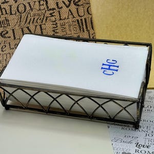 May include: A white linen napkin holder with a black metal frame. The napkins are monogrammed with the letters "CHG" in blue thread.