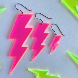 May include: Bright pink lightning bolt earrings in three sizes, with silver-colored ear wires. The earrings are made of a translucent material, and are set against a white background with neon green lightning bolt shapes.