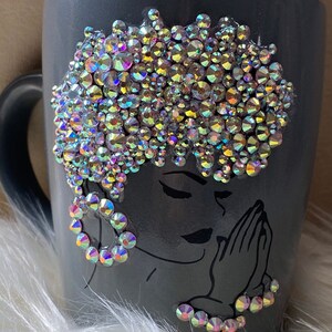May include: Gray ceramic mug with a black and white illustration of a woman with her hands clasped in prayer. The woman's hair is decorated with iridescent rhinestones.