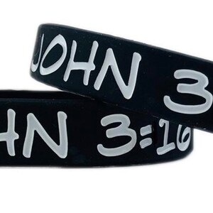 May include: Two black rubber wristbands with the text "JOHN 3:16" in white lettering. The bands are slightly curved, suggesting they are flexible and designed to fit around a wrist. The text is clearly visible against the dark background.