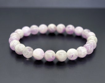 Puede incluir: Un brazalete hecho de cuentas de piedras preciosas de color púrpura claro y blanco. Las cuentas son redondas y tienen un acabado liso y pulido.