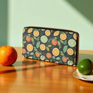 May include: A rectangular wallet with a dark background, patterned with oranges, limes, and green leaves. It has a gold-coloured zip. An orange and a lime are on a wooden table. The wallet is approximately 20 cm long.