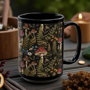 May include: Black ceramic mug featuring a detailed botanical design with mushrooms, ferns, and wildflowers. The mug has a black handle and rim, and the design is set against a dark background. The mug is likely 15 ounces.