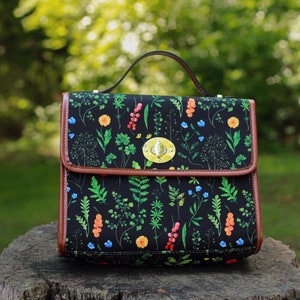 May include: A black satchel bag with a brown leather trim and a floral print featuring green leaves, red berries, and blue and yellow flowers. The bag has a top handle and a brass clasp.