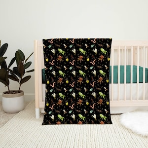 May include: A black baby blanket with a repeating pattern of colorful insects, including spiders, worms, and flies. The blanket is draped over a light-colored wooden crib with a teal green fitted sheet. A potted plant and rug are in the background.