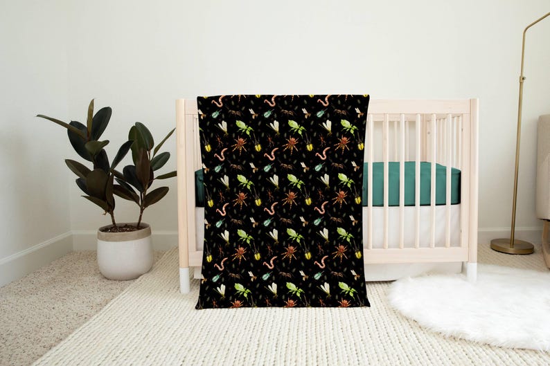 May include: A black baby blanket with a repeating pattern of colorful insects, including spiders, worms, and flies. The blanket is draped over a light-colored wooden crib with a teal green fitted sheet. A potted plant and rug are in the background.