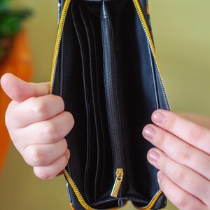 Op de afbeelding: Open zwarte portemonnee met een gele rits en een gouden ritstrekker. De portemonnee heeft meerdere compartimenten en een patroon aan de buitenkant. De portemonnee wordt opengehouden en onthult de binnenkant.