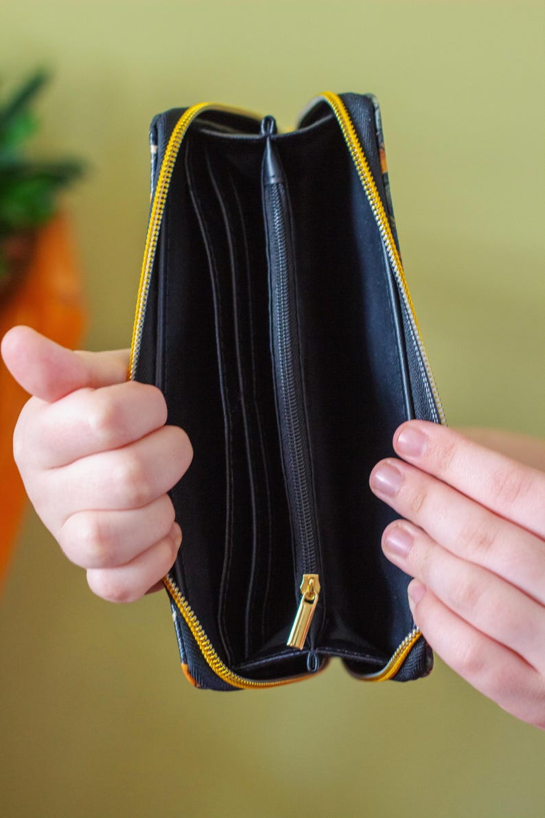 Op de afbeelding: Open zwarte portemonnee met een gele rits en een gouden ritstrekker. De portemonnee heeft meerdere compartimenten en een patroon aan de buitenkant. De portemonnee wordt opengehouden en onthult de binnenkant.