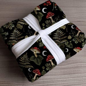May include: A black fabric with a repeating pattern of red and white mushrooms, green ferns, and crescent moons. The fabric is wrapped and tied with a white ribbon.