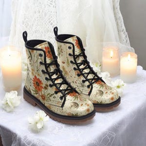 May include: A pair of cream-colored leather boots with black laces and a floral print featuring sunflowers, moons, and stars. The boots have a brown rubber sole.