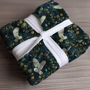 Puede incluir: Un textil plegado con un fondo verde azulado oscuro con búhos blancos, flores amarillas y follaje verde. Está envuelto y asegurado con una banda blanca gruesa. El artículo es probablemente una manta o un textil similar.