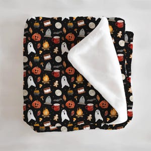 May include: A black Halloween-themed blanket with a repeating pattern of pumpkins, ghosts, campfires, tombstones, and mugs. The blanket has a white, fluffy underside. The design includes a full moon, bats, and gingerbread men.