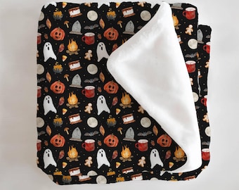 May include: A black Halloween-themed blanket with a repeating pattern of pumpkins, ghosts, campfires, tombstones, and mugs. The blanket has a white, fluffy underside. The design includes a full moon, bats, and gingerbread men.