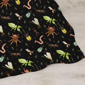 May include: A black blanket featuring a repeating pattern of various insects, including spiders, ants, beetles, and dragonflies. The insects are in shades of green, yellow, brown, and orange, with a watercolor style. The blanket is draped over a light wood surface.