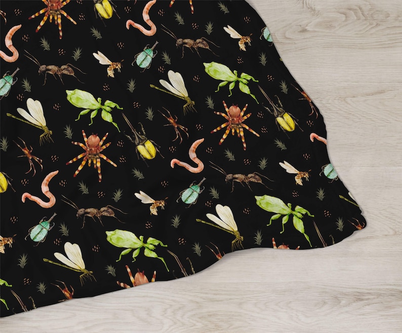 May include: A black blanket featuring a repeating pattern of various insects, including spiders, ants, beetles, and dragonflies. The insects are in shades of green, yellow, brown, and orange, with a watercolor style. The blanket is draped over a light wood surface.