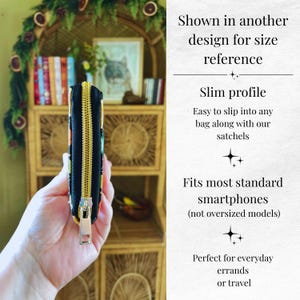 Op de afbeelding: Zwarte portemonnee met een gouden rits, vastgehouden in een hand. De portemonnee heeft een slank profiel en past op de meeste standaard smartphones. Tekst: "Getoond in een ander ontwerp ter referentie van de maat".
