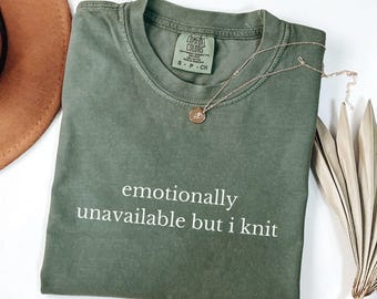 May include: Moss green t-shirt with the text "emotionally unavailable but i knit" in white. A gold necklace with a pendant rests on the shirt. The shirt is folded and placed on a white surface.