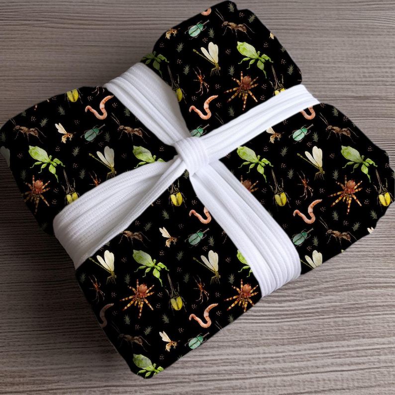 May include: A black fabric with a repeating pattern of insects, including spiders, ants, and dragonflies, is wrapped and tied with a white ribbon. The insects are in shades of green, brown, and yellow. The fabric is on a wooden surface.