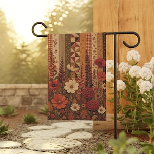 Peut inclure: Un drapeau de jardin rectangulaire présentant un motif patchwork vertical de rayures et de formes géométriques dans des tons automnaux chauds. La partie inférieure affiche un arrangement dense de fleurs rouges, orange et blanches.