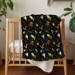 May include: A black baby blanket featuring a repeating pattern of colorful insects, including spiders, dragonflies, and worms. The blanket is draped over a wooden crib, with a white, fluffy underside visible.