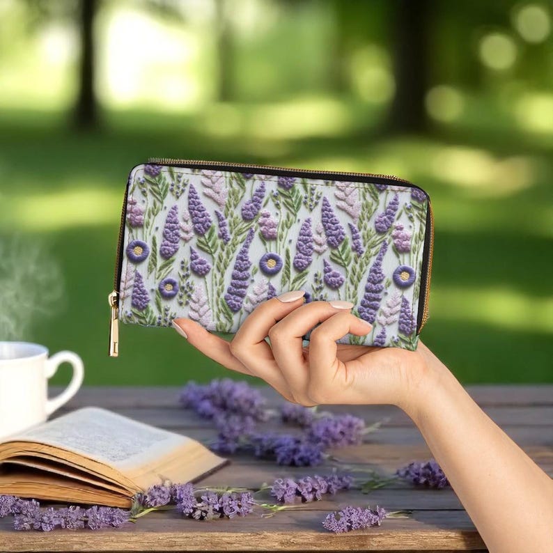 Op de afbeelding: Een portemonnee met ritssluiting met een lavendelbloemmotief. De portemonnee heeft een lichtblauwe achtergrond met paarse lavendelbloemen en groene bladeren. De rits is goudkleurig.