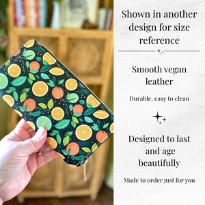 Op de afbeelding: Een zwarte vegan leren portemonnee met een levendig citrusfruitpatroon. Het ontwerp toont sinaasappel-, limoen- en citroenschijfjes met groene bladeren. De tekst zegt "Smooth vegan leather" en "Durable, easy to clean".