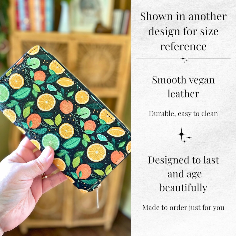 Op de afbeelding: Een zwarte vegan leren portemonnee met een levendig citrusfruitpatroon. Het ontwerp toont sinaasappel-, limoen- en citroenschijfjes met groene bladeren. De tekst zegt "Smooth vegan leather" en "Durable, easy to clean".